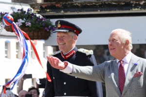 Two elderly men in a parade; a uniformed officer with a red-trimmed hat and a gray-suited man waving colorful ribbons.