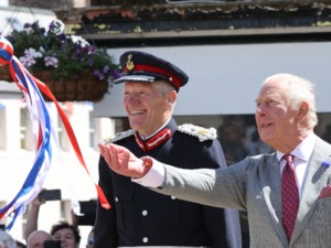 Two elderly men in a parade; a uniformed officer with a red-trimmed hat and a gray-suited man waving colorful ribbons.
