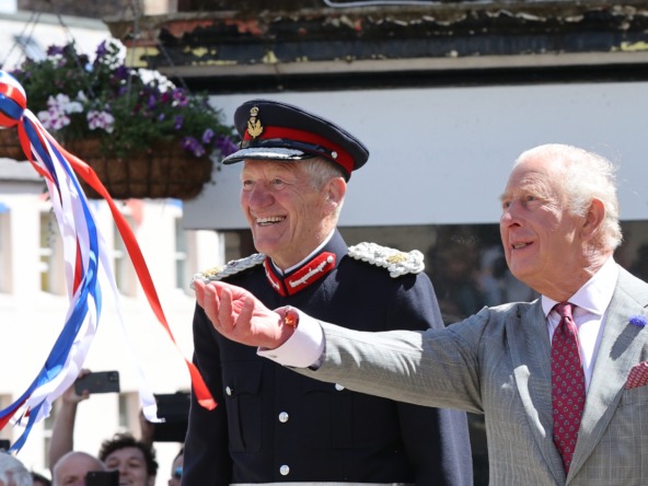 Two elderly men in a parade; a uniformed officer with a red-trimmed hat and a gray-suited man waving colorful ribbons.