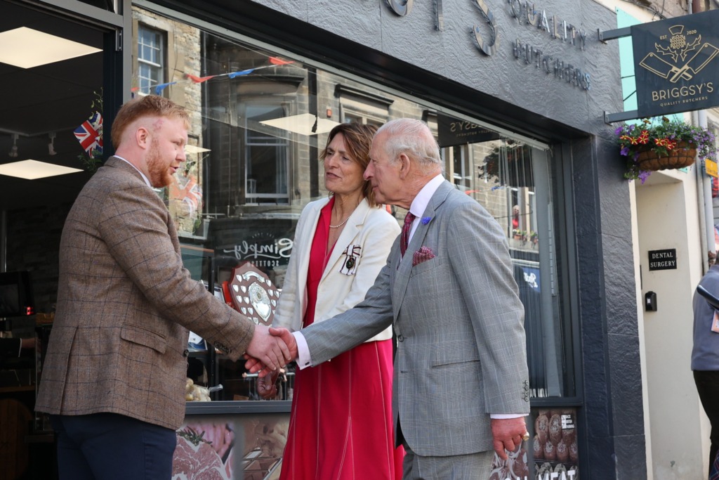 Three adults in formal attire shake hands outside a shop window, with a man in a brown plaid jacket, a woman in a red dress, and an older man in a gray suit; storefront signage and hanging flower basket visible.