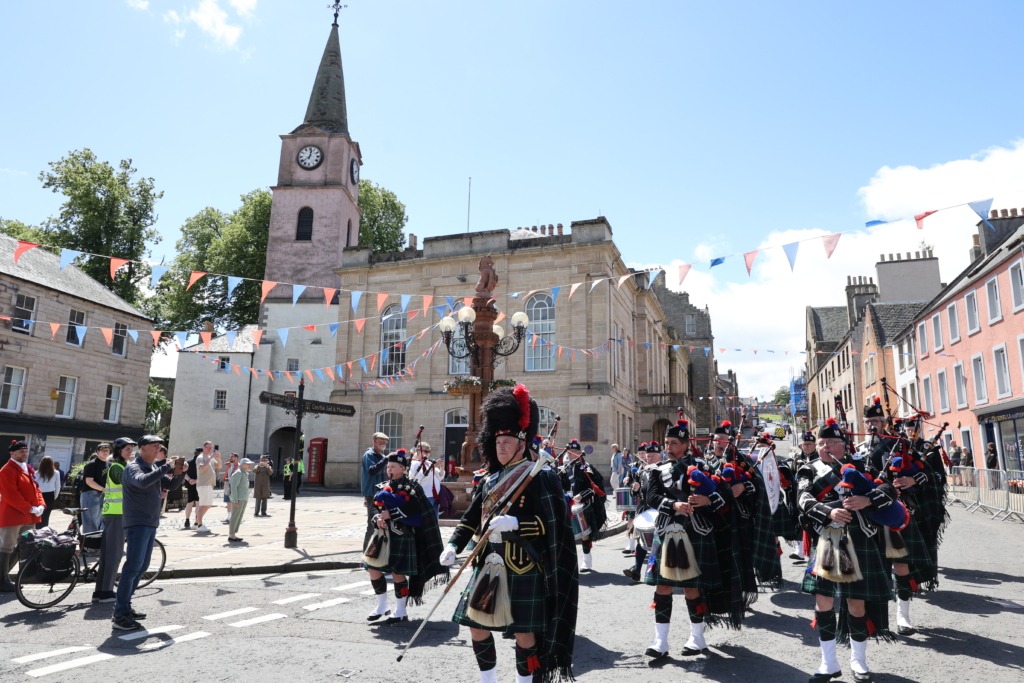 Parade with bagpipers in uniforms marching down a sunny street, bunting and historic buildings overhead.