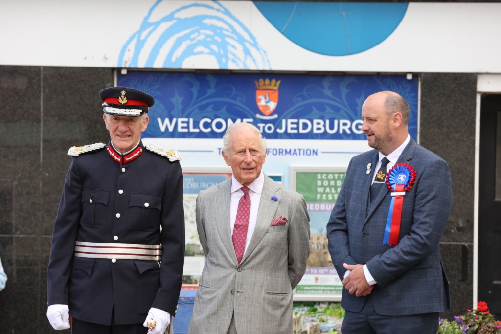 Three men in formal attire pose in front of a blue 'Welcome to Jedburgh' banner (ceremonial event).