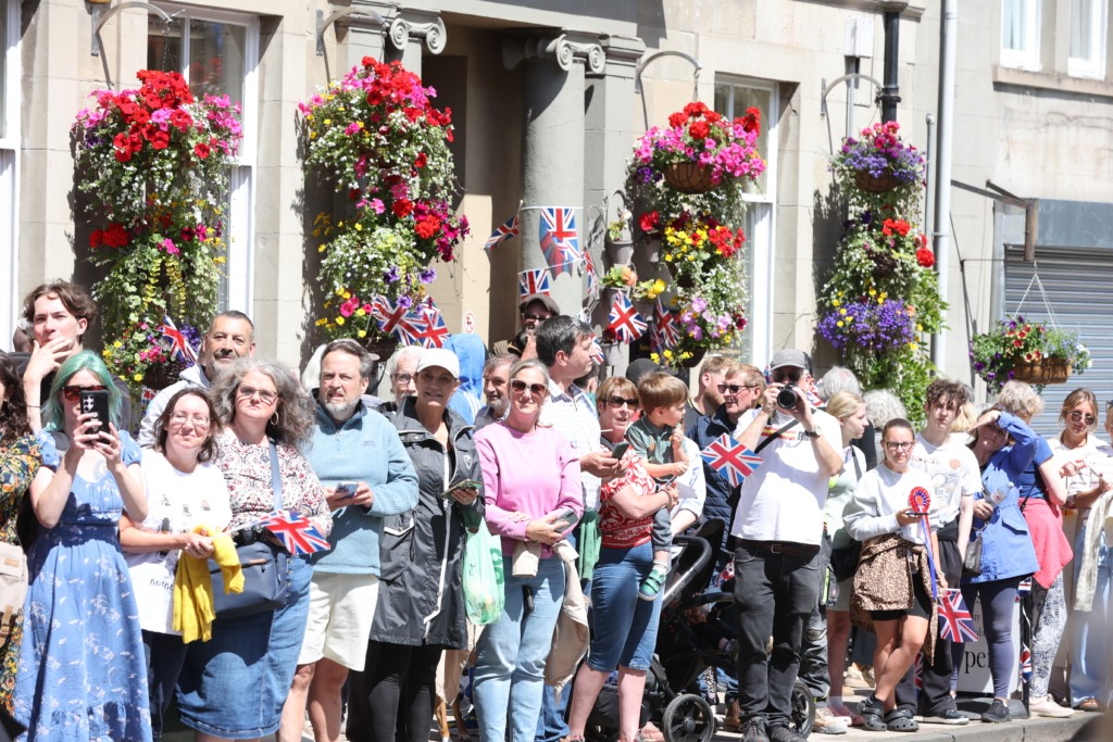 Crowd of people on a sunny street during a celebration, holding Union Jack flags amid hanging flower baskets in the background.
