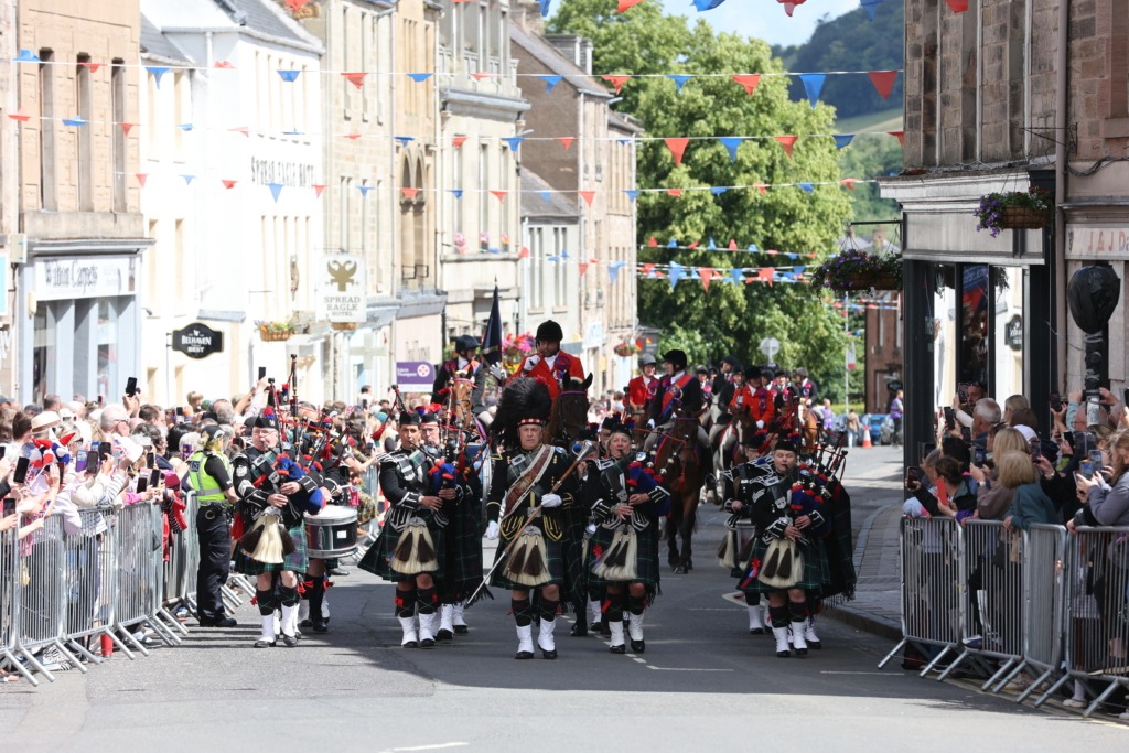 Parade on a town street with bagpipers in kilts marching between crowd barriers and colorful bunting overhead.