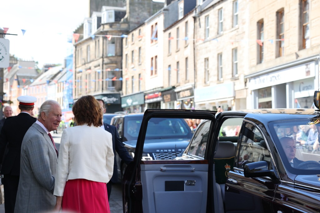 Two formally dressed people stand near a black Range Rover with its rear door open on a busy street, brick buildings overhead bunting.”]}{