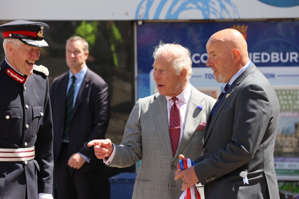 Three men in formal attire outdoors, one in a military-style dress uniform, smiling and talking together.