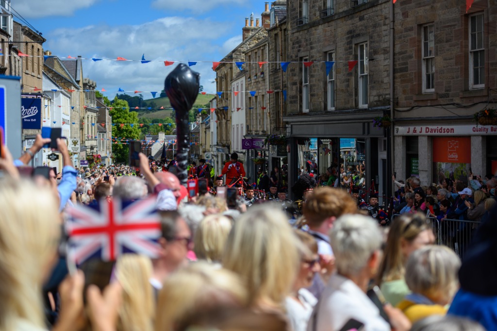 Crowded town street during a parade with red and blue bunting and Union Jack flags overhead, spectators on both sides.