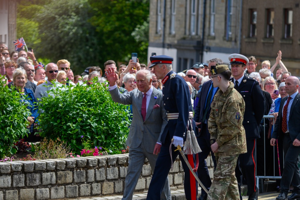 Crowd watches a parade as uniformed military personnel walk along a street, with a man in a suit waving at the foreground and spectators behind a stone wall.