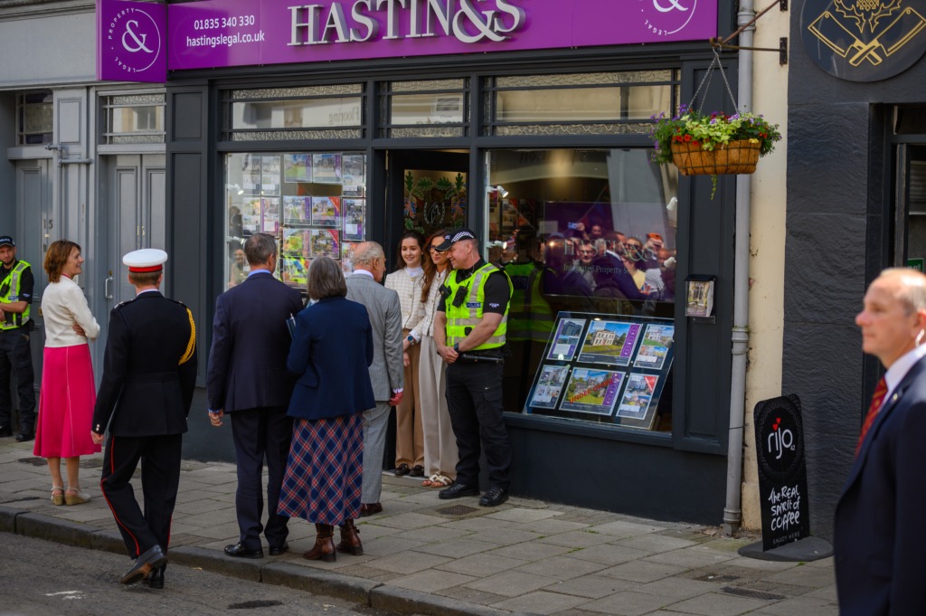 Group of civilians and police officers stand outside a shop with a purple Hastings sign and colorful window displays, near a hanging flower basket.