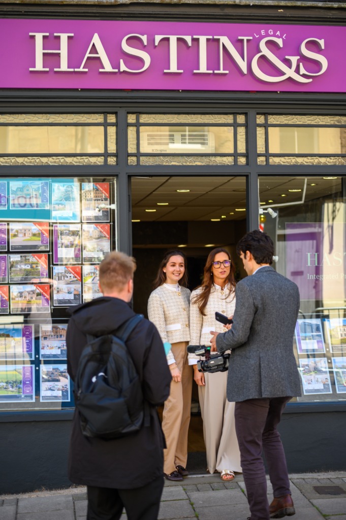 Group of people chat outside a real estate agency storefront with a large purple sign above and window posters displaying properties.