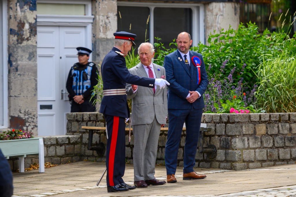 Man in ceremonial military uniform shakes hands with an older gentleman in a gray suit during an outdoor event, with another suited man and a police officer in the background.
