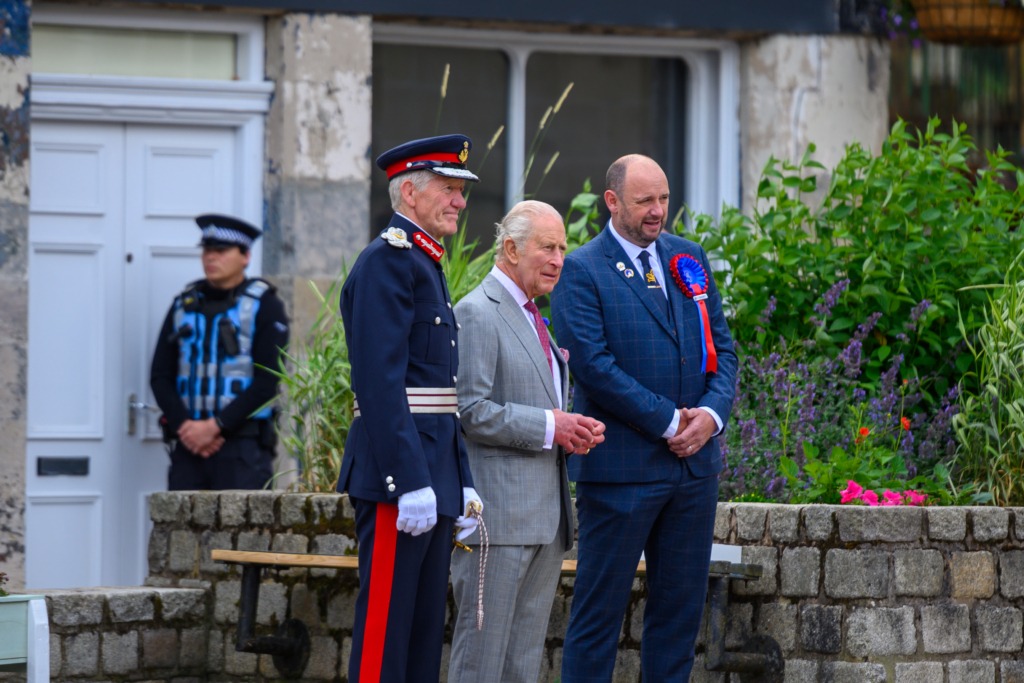 Three men stand outdoors: two in ceremonial uniforms and one in a gray suit, near a stone wall and flowering shrubs.